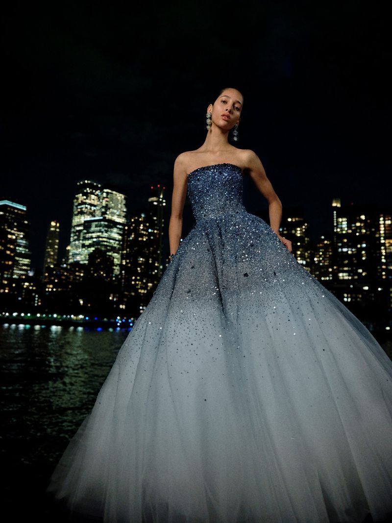 Woman in a shimmering blue gown poses against a dark city skyline with reflections on water, creating a high-fashion editorial scene.