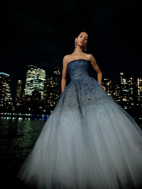 Woman in a shimmering blue gown poses against a dark city skyline with reflections on water, creating a high-fashion editorial scene.