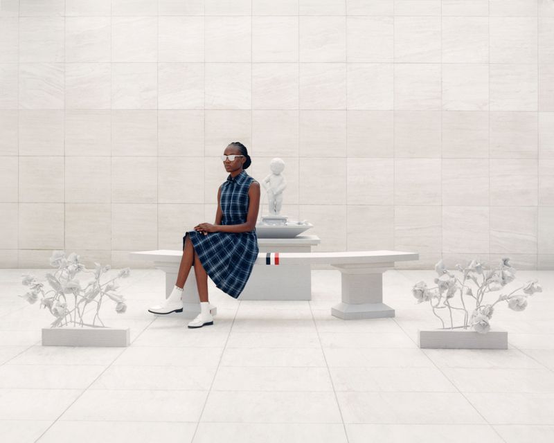 Adult woman in stylish blue plaid dress sitting on white bench with sculptural accents in a bright, minimalist indoor setting.