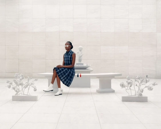 Adult woman in stylish blue plaid dress sitting on white bench with sculptural accents in a bright, minimalist indoor setting.