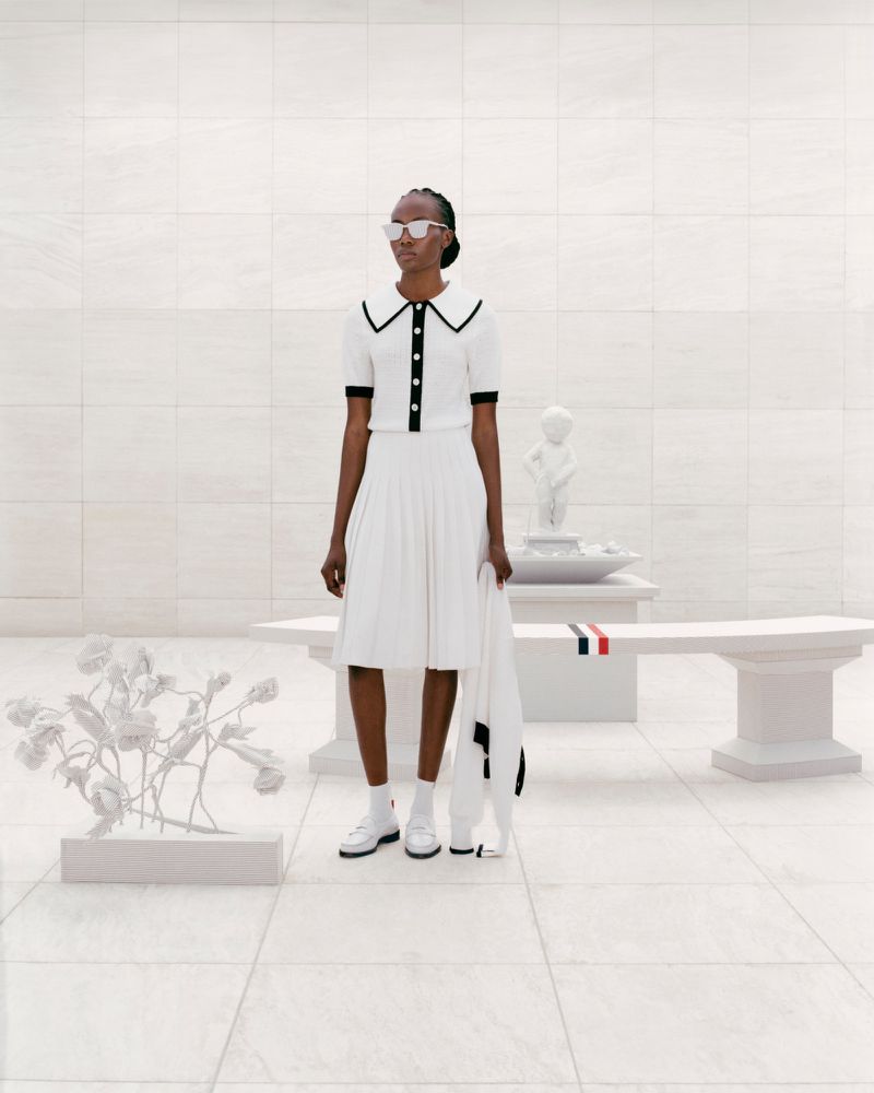 Woman poses in a white, collared dress and sunglasses in a bright white architectural space, standing next to a white bench and sculpture.