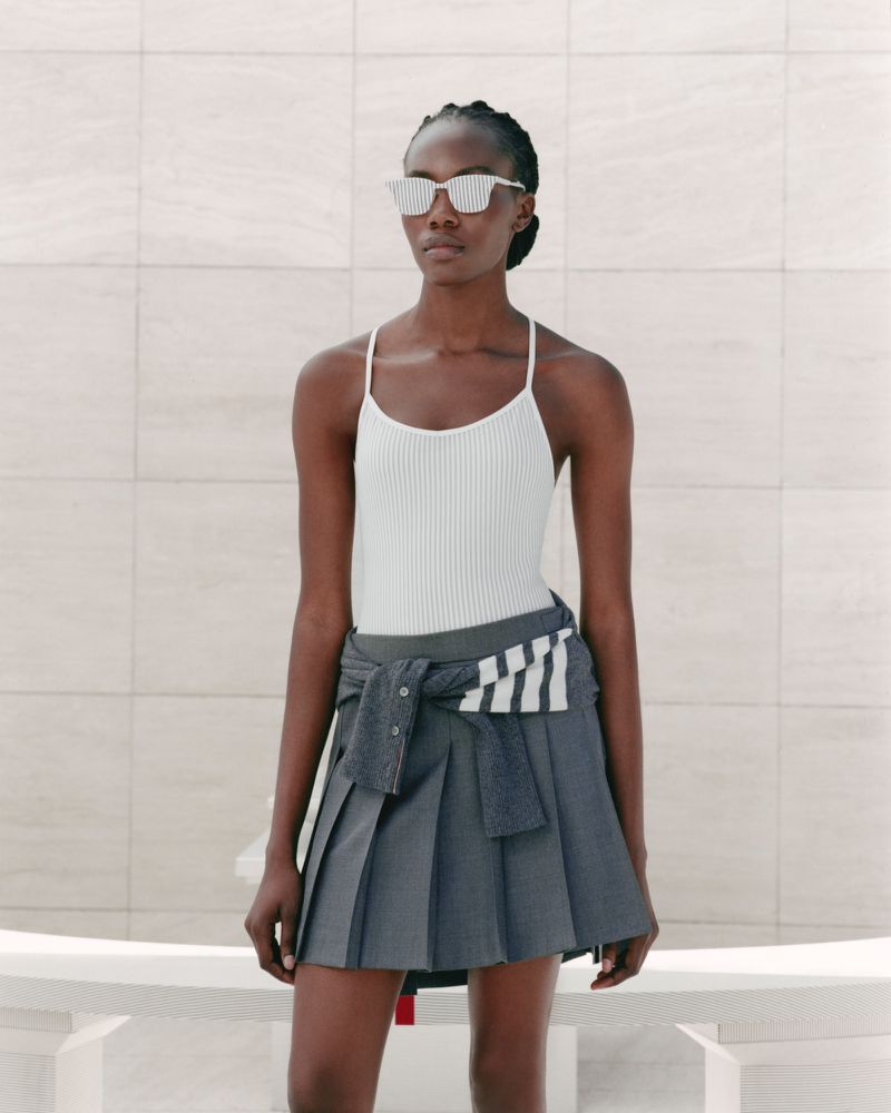 A black female fashion model poses against a tiled wall wearing sunglasses, a white top, and a pleated gray skirt with sweater around her waist.