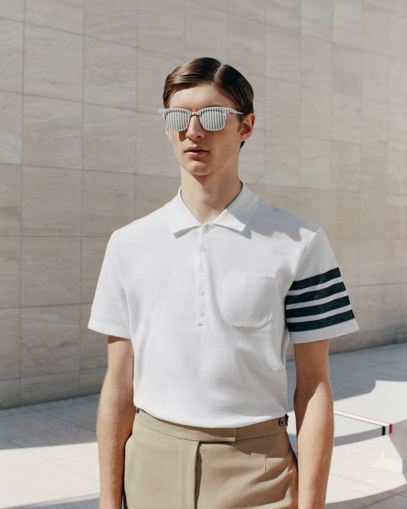 A young adult male wearing a white polo shirt, khaki pants, and striped sunglasses stands against a light-colored wall in natural light.