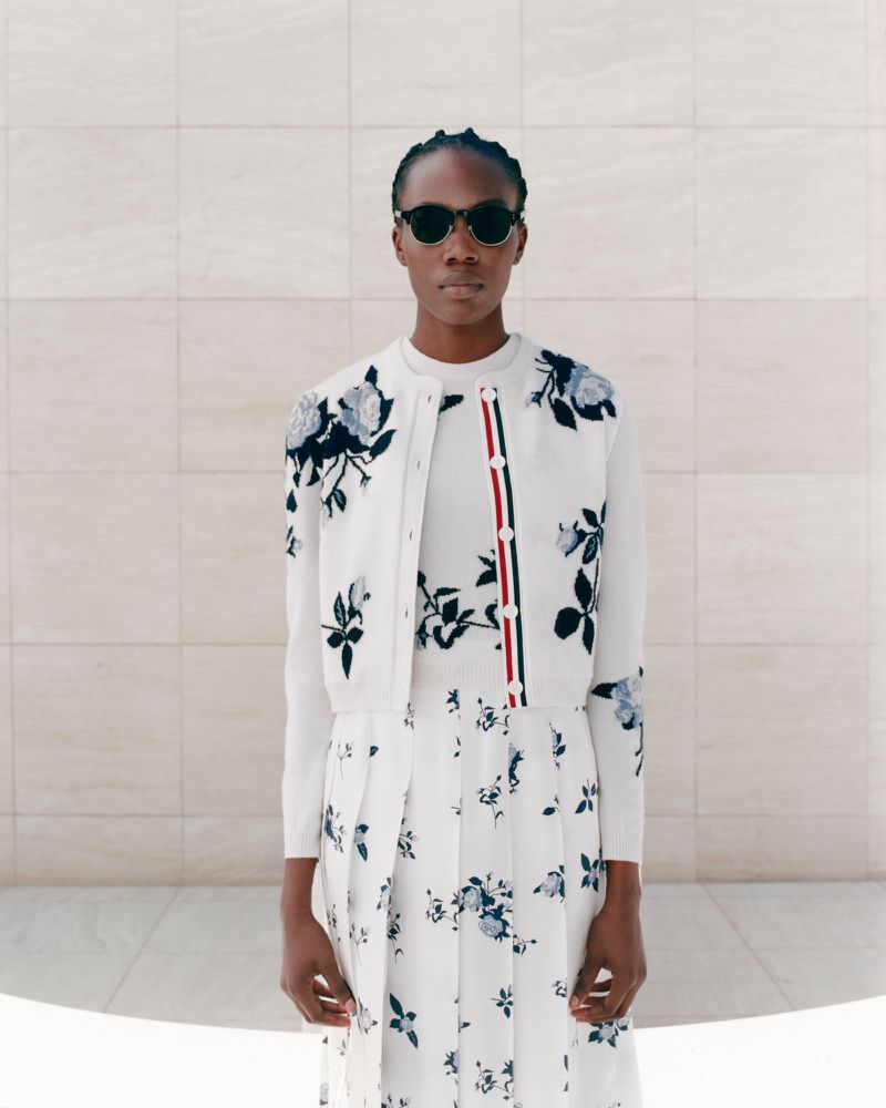 A woman wearing sunglasses, a white floral cardigan, top, and pleated skirt poses in front of a cream-colored tiled wall and floor.