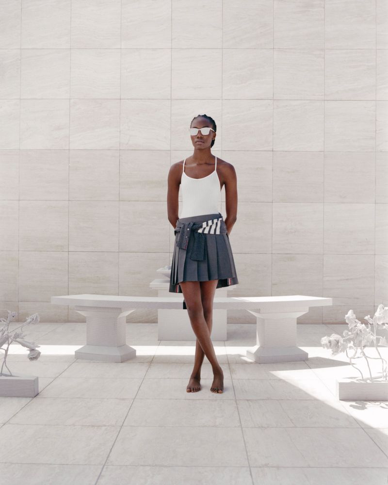 Fashion shot of a barefoot Black woman in sunglasses, tank top, and pleated skirt, posing on a sunlit white stone patio near a bench.