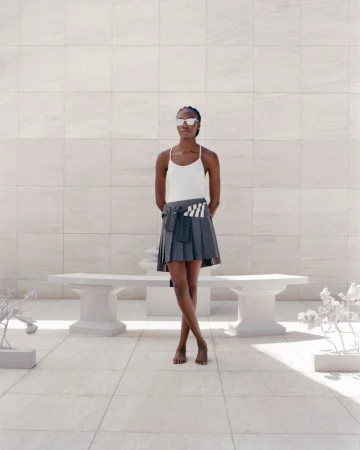 Fashion shot of a barefoot Black woman in sunglasses, tank top, and pleated skirt, posing on a sunlit white stone patio near a bench.
