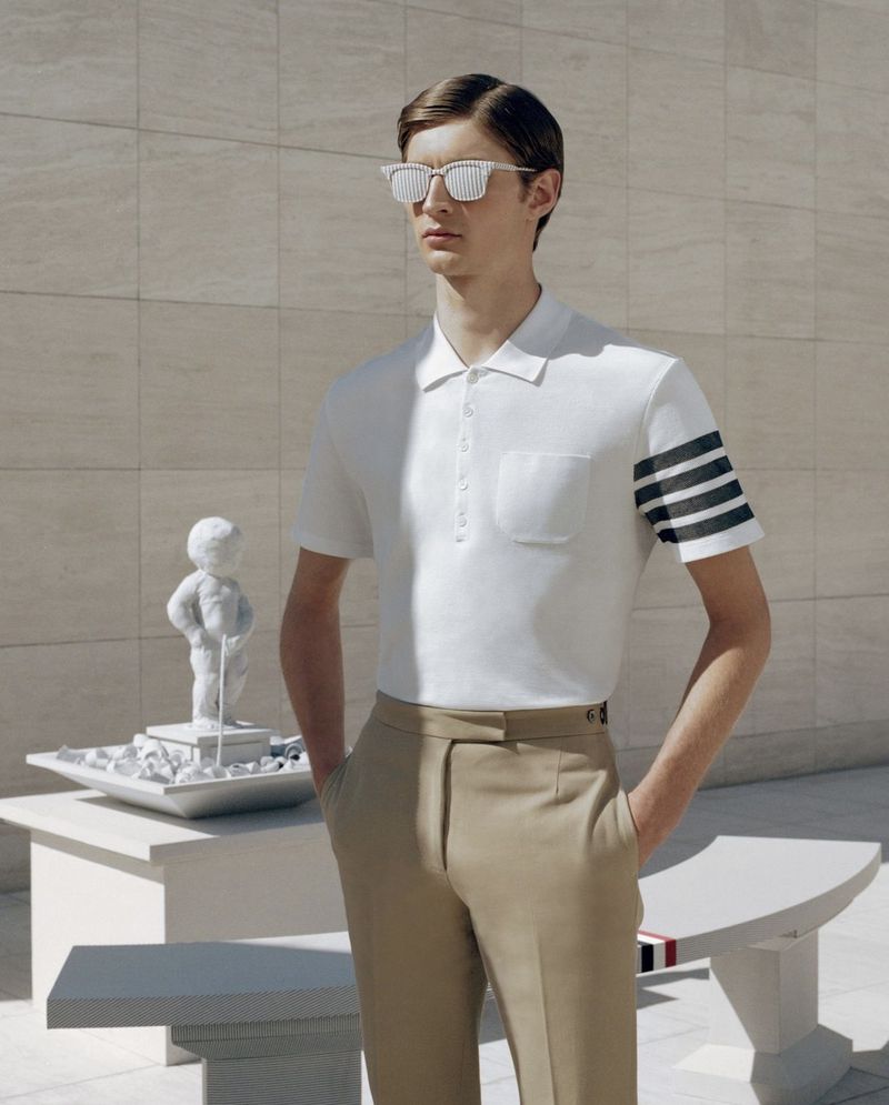 An adult male model wears a white polo shirt with khaki pants while posing next to a statue in an outdoor setting on a sunny day.