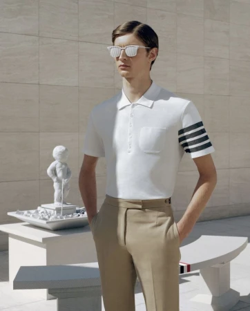 An adult male model wears a white polo shirt with khaki pants while posing next to a statue in an outdoor setting on a sunny day.