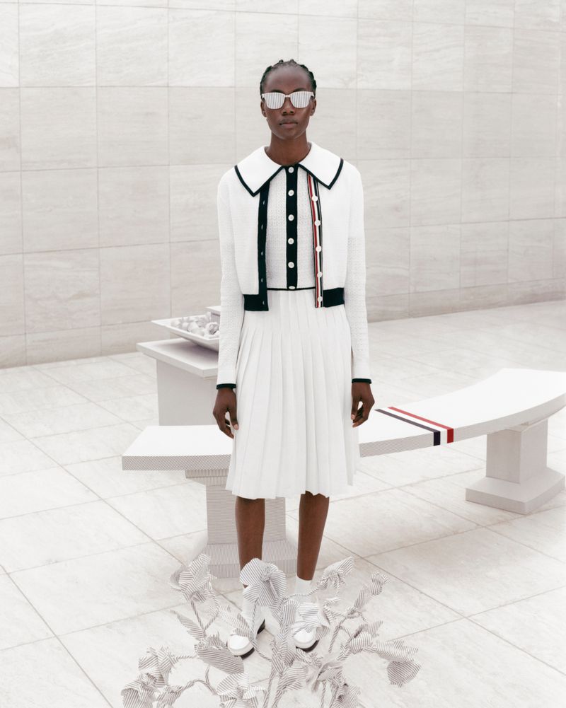 Elegant African American woman in a white pleated dress and cardigan poses indoors with modern sculpture and geometric background.