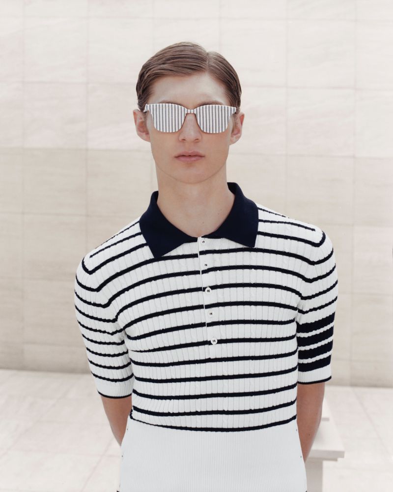 Striking studio shot of a slender adult male model with blond hair wearing striped sunglasses and a navy and white striped polo shirt.