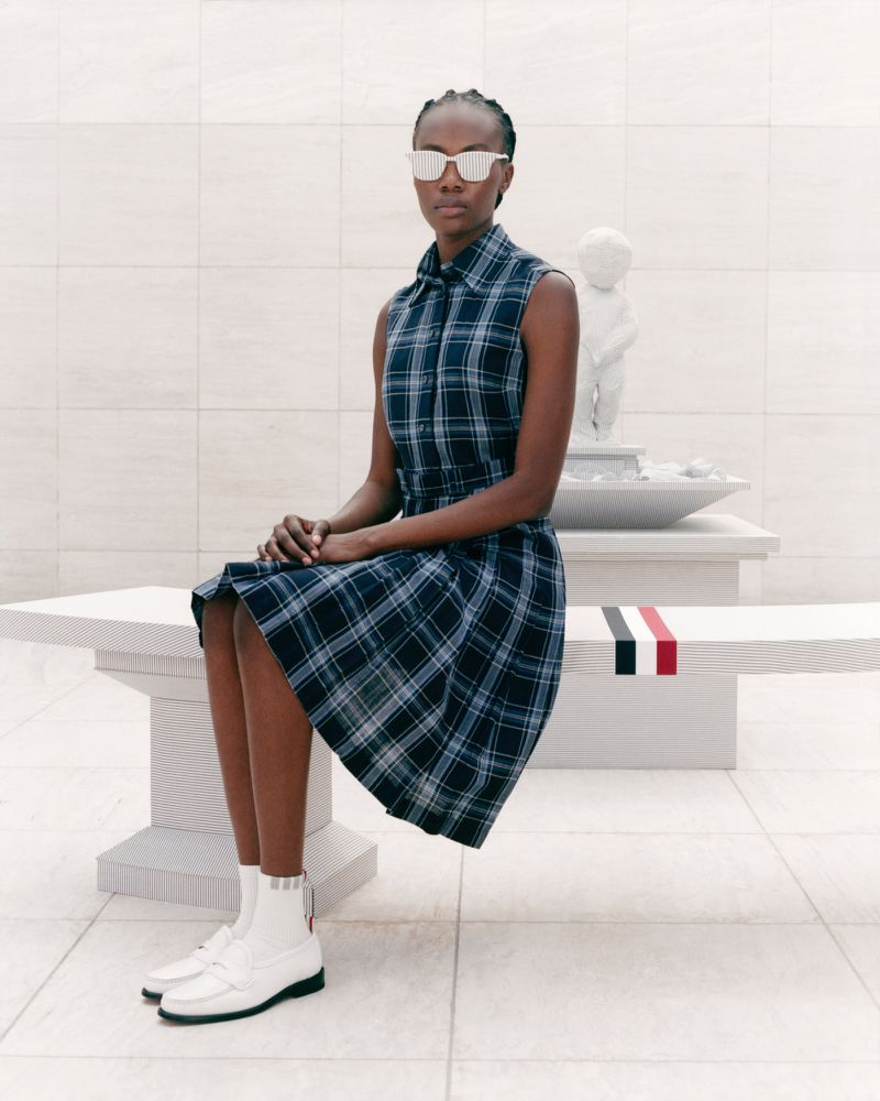 Adult female model in plaid dress and sunglasses sits on a bench in bright, modern setting near a small sculpture, showcasing a fashion style.