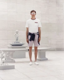 Model Robbie Lyons stands in a white t-shirt, tailored shorts, tied navy sweater, and white sneakers before a stone courtyard backdrop.