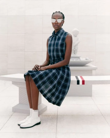 Model Robbie Lyons seated on a bench wearing a blue plaid sleeveless dress, white loafers, and sunglasses for Thom Browne S/S 24.