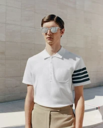 Male model wearing sunglasses and a white four-bar stripe polo shirt paired with beige trousers against a stone wall.