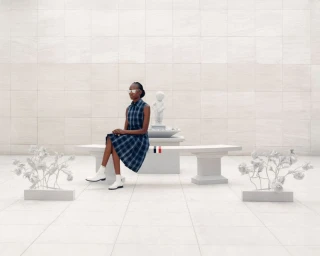 Model wearing a blue plaid dress and white boots seated on a stone bench in a monochrome set by Thom Browne