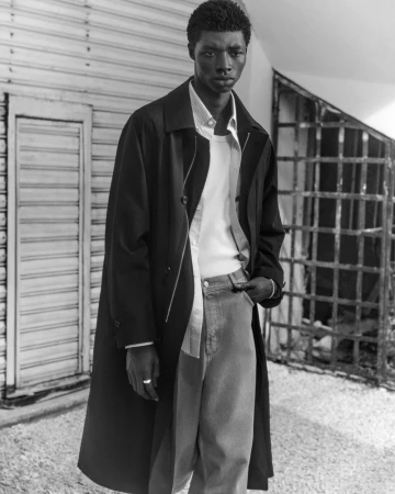Black and white portrait of an African American man in stylish outerwear and jeans standing outdoors with a neutral facial expression.
