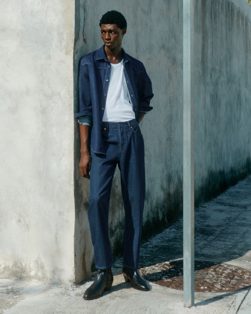 An African American man poses against a concrete wall wearing a white t-shirt, a blue denim shirt and pants, and black boots in natural light.