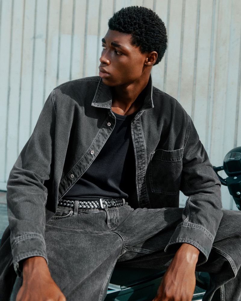 A fashionable Black man in a grey denim jacket and jeans sits on a motorcycle outdoors, looking to the side with a serious expression.