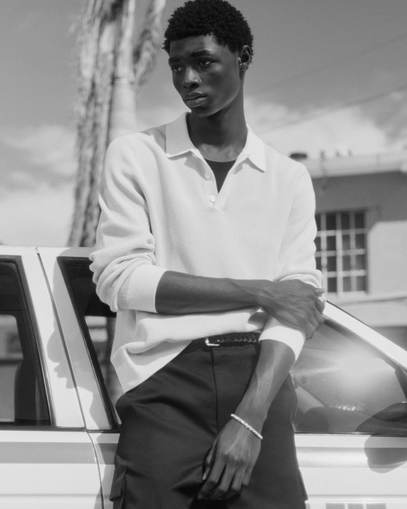 Black and white portrait of a Black male model wearing a polo shirt and leaning on a vintage car with a palm tree background.
