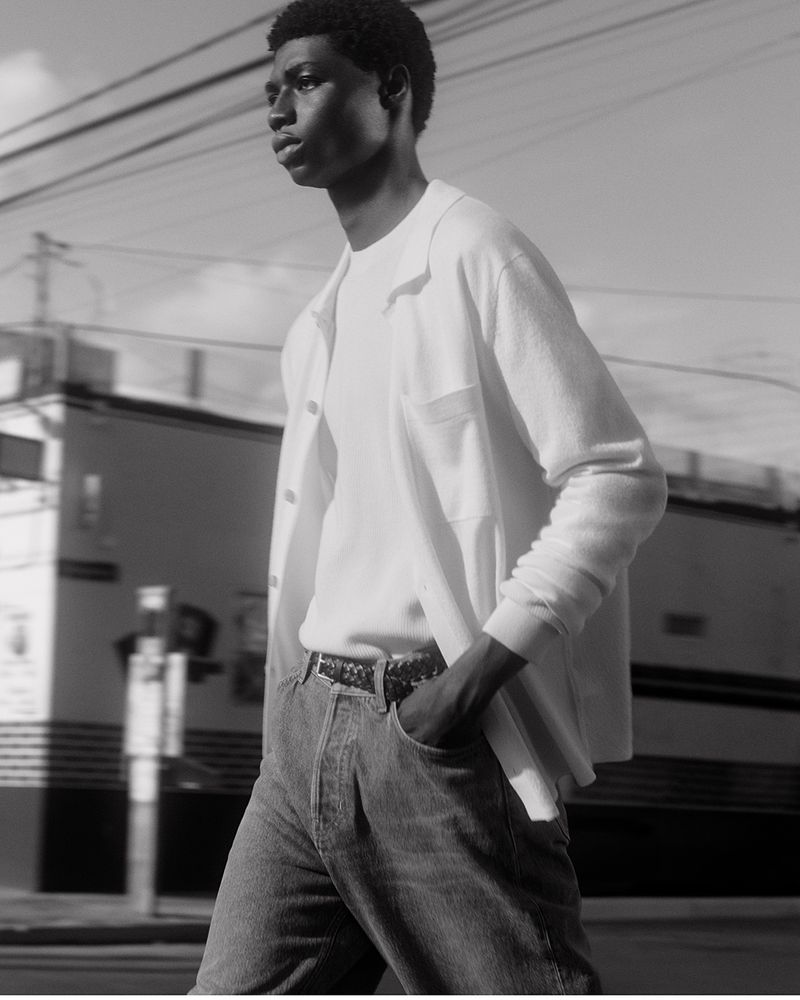 Monochrome full shot of a Black male model in white shirt and blue jeans walking on a city street with buildings and wires in the background.
