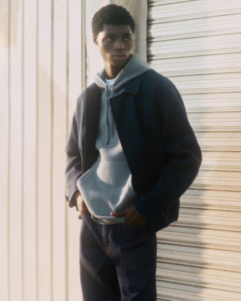 A young Black male model poses outdoors wearing a gray hooded sweatshirt, blue jacket, and dark blue jeans in a fashion editorial shot.