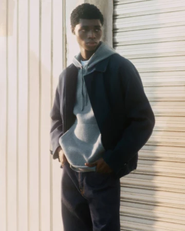 A young Black male model poses outdoors wearing a gray hooded sweatshirt, blue jacket, and dark blue jeans in a fashion editorial shot.