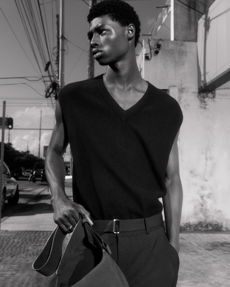 Black and white portrait of an African American male model in a vest and trousers, holding a bag, against an urban background.