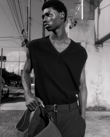 Black and white portrait of an African American male model in a vest and trousers, holding a bag, against an urban background.