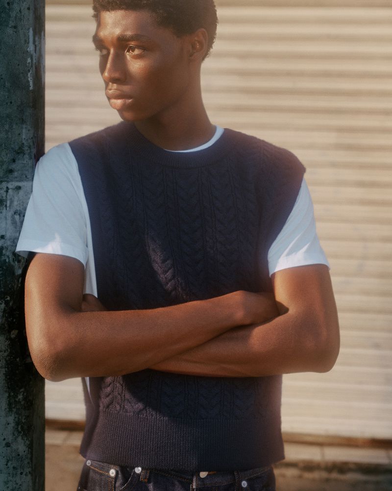 A young Black man poses outdoors in a navy sweater vest, white t-shirt, and blue jeans, with crossed arms and a serious expression.
