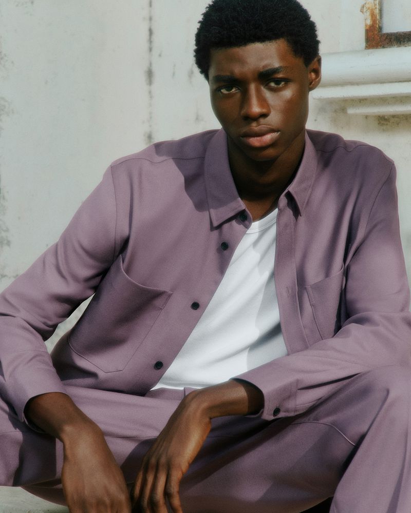 Portrait of a dark-skinned man sitting outdoors wearing a lavender button-down shirt over a white t-shirt and matching pants.