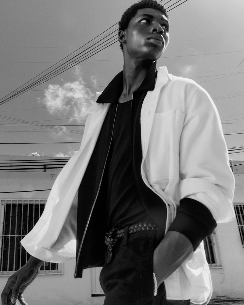 Monochrome fashion shot of a Black man in a white jacket over a black top, set against a building and sky backdrop.
