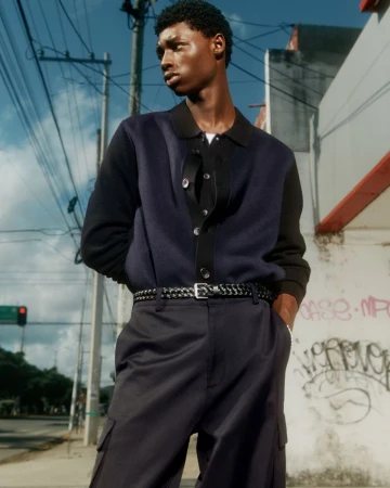 Dark-skinned man in a navy cardigan and dark trousers stands outside with graffiti in the background, captured in natural light.