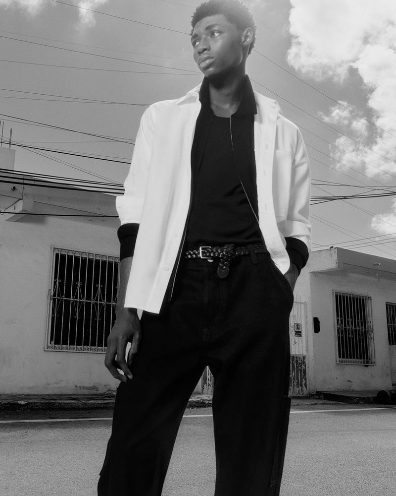 Black and white portrait of an African American male outdoors wearing casual clothes with a building and cloudy sky in the background.