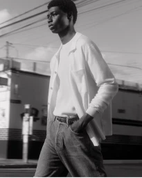 Black man wearing a white buttoned shirt and jeans outdoors against a building and overhead utility lines