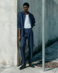 Full-length portrait of a black man wearing a dark denim shirt, white t-shirt, and jeans leaning against a concrete wall