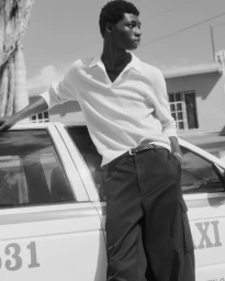 Black male model leaning against a white taxi wearing a collared polo shirt and dark trousers for COS Spring 2024 prelude campaign.