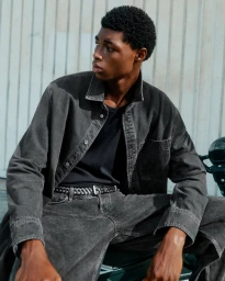 Zaram Obasi seated on a motorcycle wearing a washed black denim jacket and jeans against a corrugated wall.