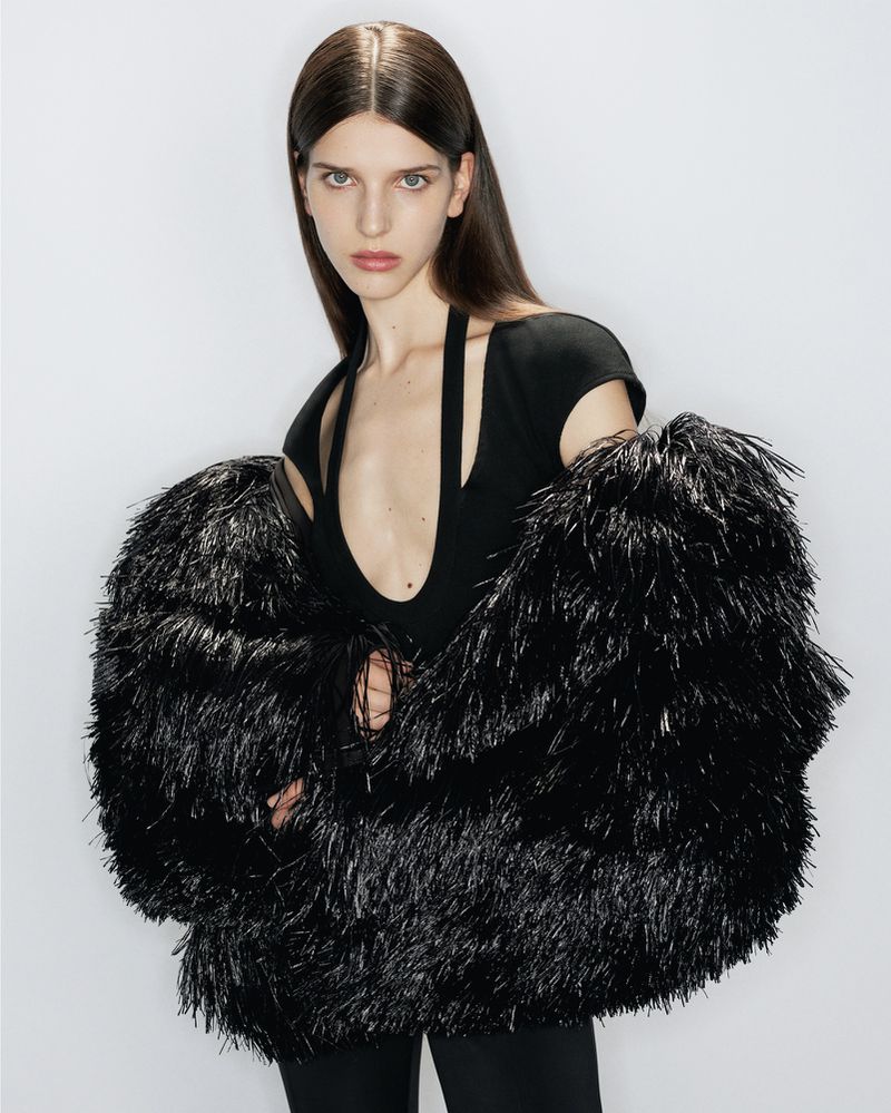 A model wearing a black top and black pants is posing in a studio with a large furry black coat over their shoulders.