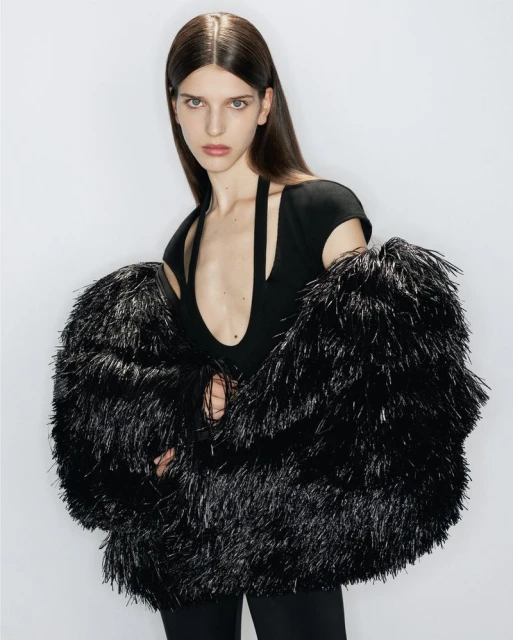 A model wearing a black top and black pants is posing in a studio with a large furry black coat over their shoulders.