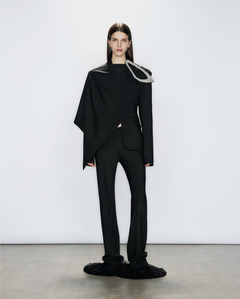 Fashion shot of a woman in a black suit with an asymmetrical shawl and unusual black shoes against a bright white backdrop.