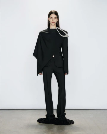 A woman standing in an asymmetrical black jacket with a silver shoulder brooch and black trousers against a white studio backdrop.