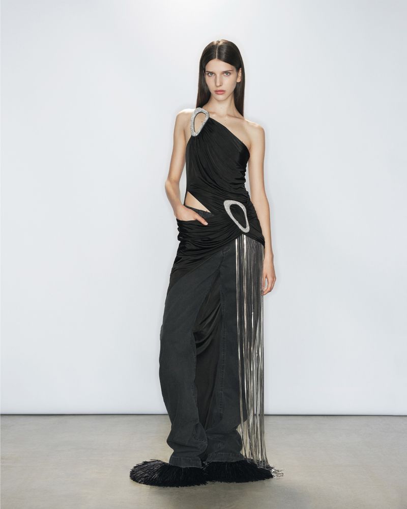 Woman poses in a black asymmetrical draped dress with fringe details against a white backdrop, full shot, fashion, studio lighting.