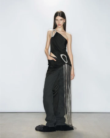 Woman poses in a black asymmetrical draped dress with fringe details against a white backdrop, full shot, fashion, studio lighting.