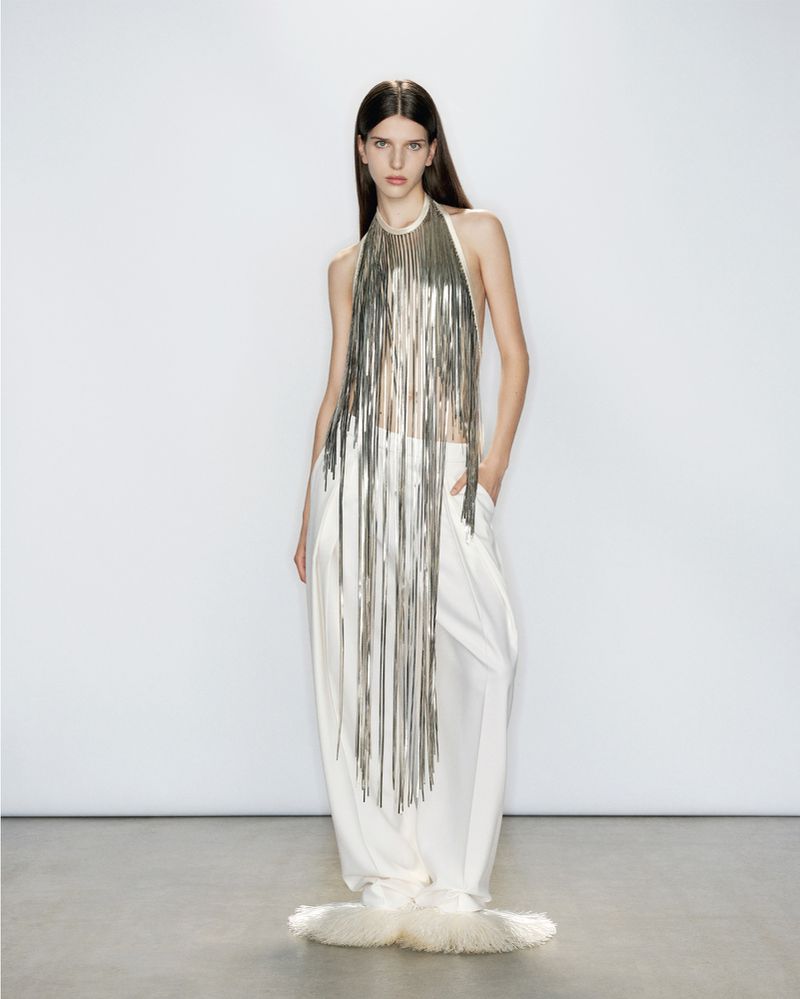 A female fashion model poses in a studio wearing a silver, fringed halter top and wide-legged white pants, standing against a white backdrop.