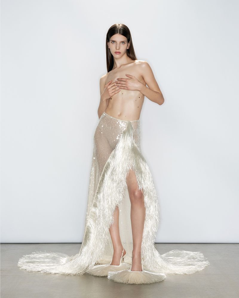 A slender woman poses in a light skirt and high heels against a seamless white backdrop in a fashion or editorial studio photograph.