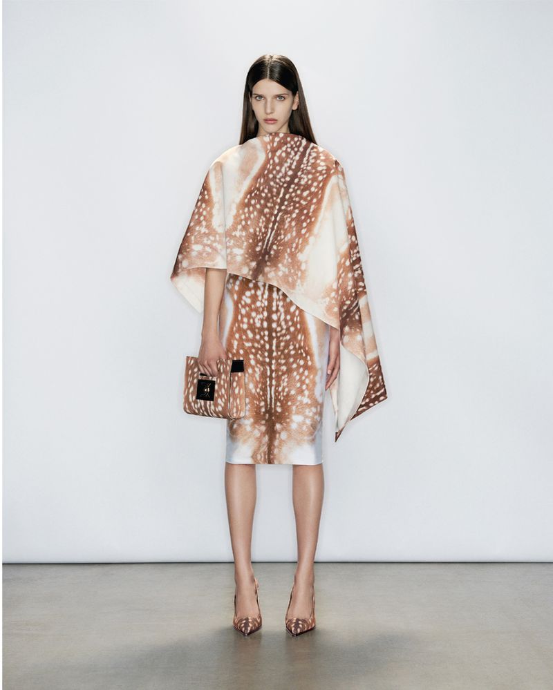 A woman poses in a deer-print dress, cape, and heels against a white backdrop, holding a matching purse in a fashion studio setting.