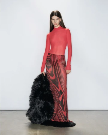 Model Eva Pujatti in a red long-sleeve top and printed red and black maxi skirt holding a black fringe wrap against a white backdrop.