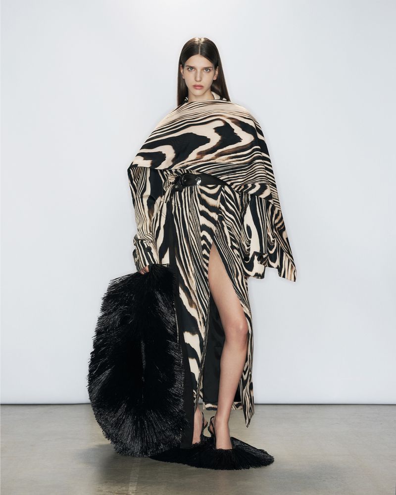 A woman is posing in a studio wearing a zebra-striped dress and carrying a large feathered bag. The backdrop is a bright white.