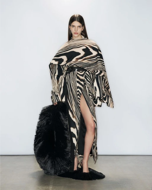 A woman is posing in a studio wearing a zebra-striped dress and carrying a large feathered bag. The backdrop is a bright white.
