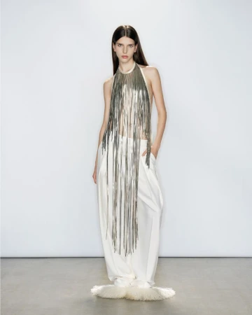 Model Eva Pujatti in a long metallic fringe halter top and wide-leg white trousers posing against a clean white studio backdrop.